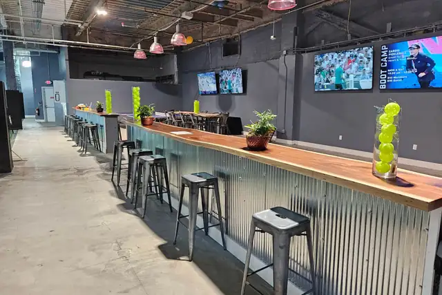 A long bar with a wooden top and corrugated metal front, lined with metal stools, under pink pendant lights and wall-mounted TVs.