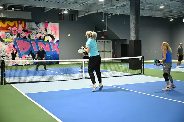 A doubles game on a blue court beside the DINK mural, a player in a light blue shirt stepping toward the ball.