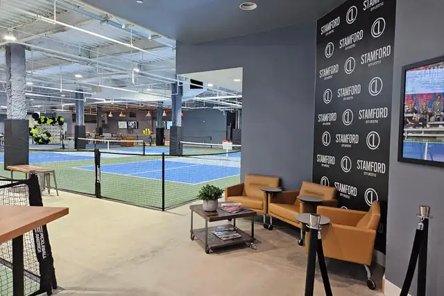 A seating corner with tan leather armchairs and a low table beside a Stamford branded backdrop, with blue courts behind a black barrier.