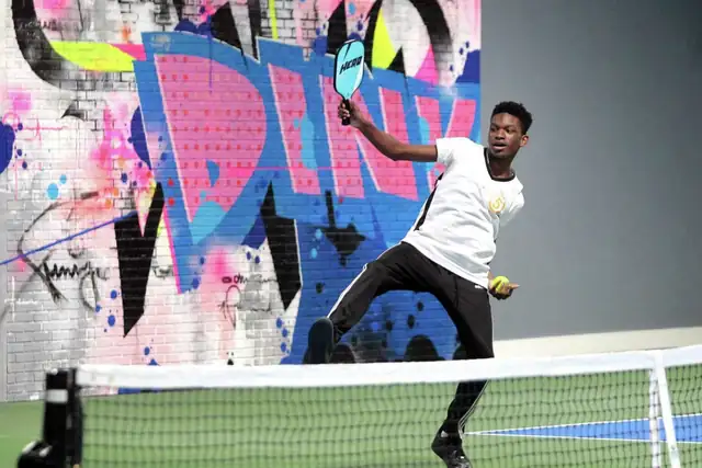 A player in a white T-shirt lunges for a shot at the net, paddle raised, in front of a pink and blue graffiti mural spelling DINK.