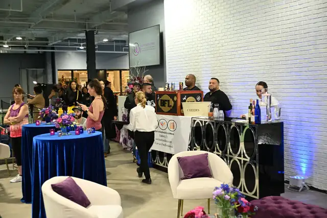 An evening event beside a white brick wall: guests mingle around tall tables draped in blue velvet, with white armchairs and a staffed bar.