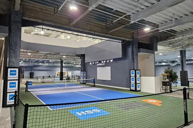 An empty blue pickleball court with green run-off, fenced by low black netting and HSS Stamford boards, with more courts beyond under an exposed ceiling.