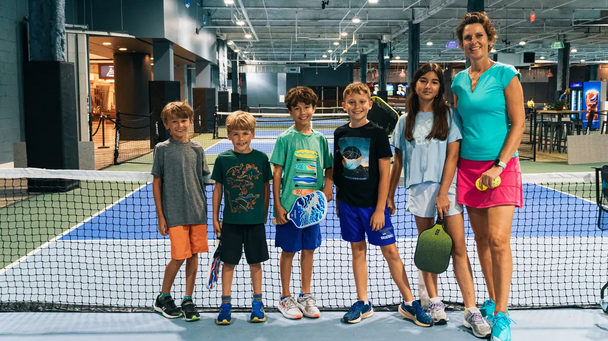 Six junior players stand in a row along the net on an indoor pickleball court at Pickleball America, holding paddles and balls, with their coach beside them.