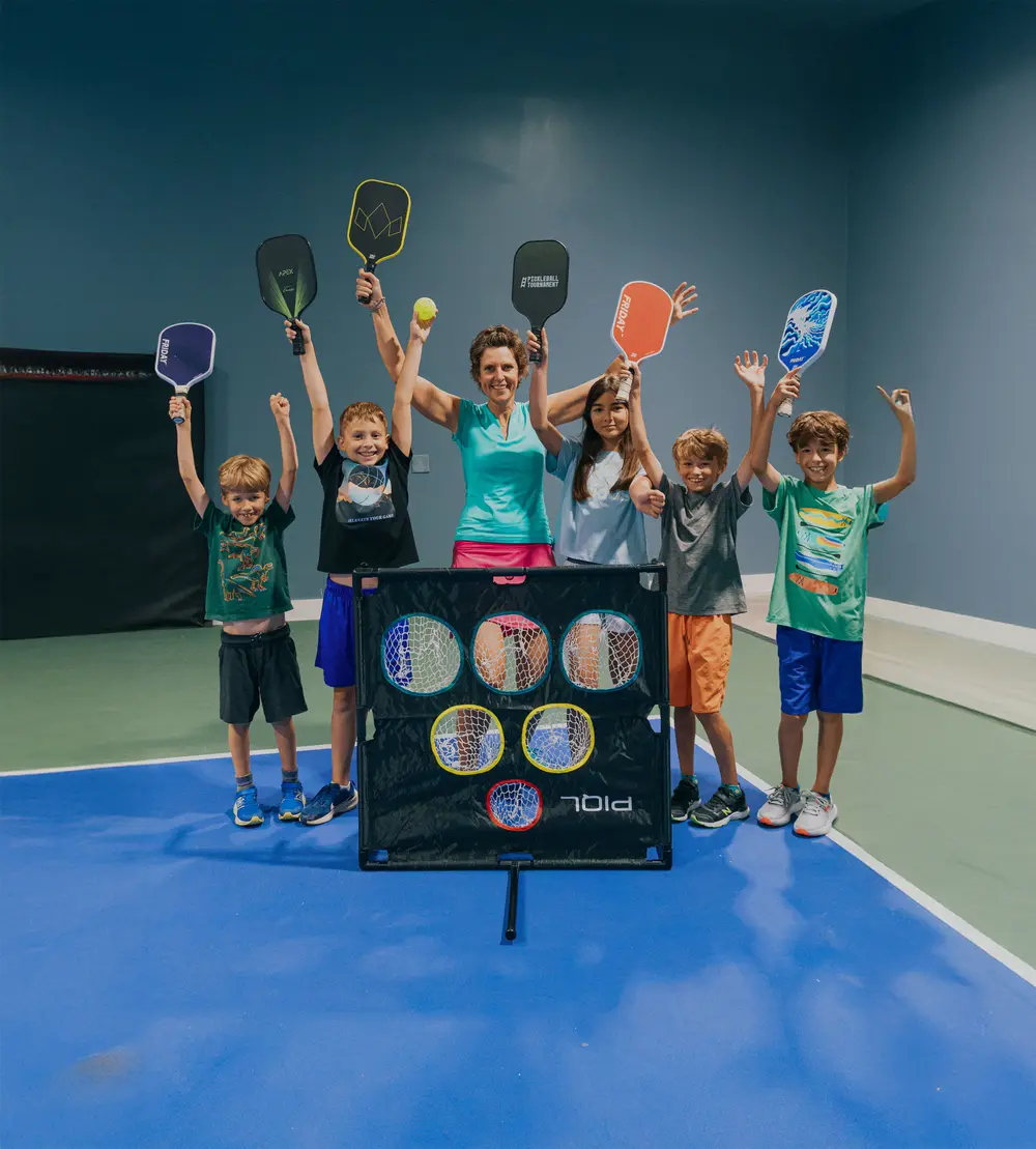 Junior players and their coach raise pickleball paddles above their heads, cheering behind a target net on an indoor court.