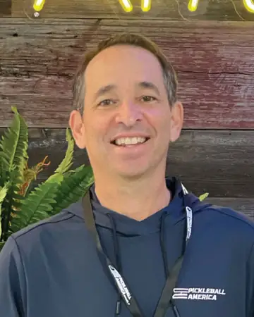 Bret Herman, smiling, in a navy Pickleball America hoodie and lanyard, in front of a wood-plank wall.