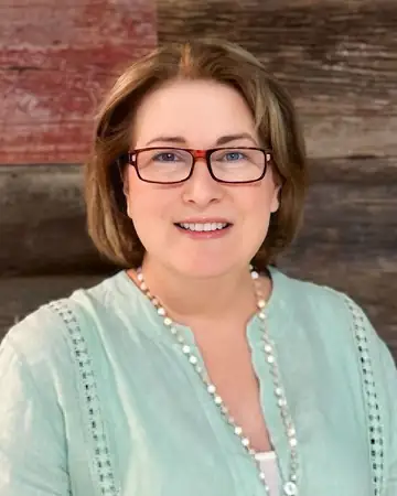 Dawn Schneider, smiling, in red-framed glasses and a mint green top, in front of a wood-plank wall.