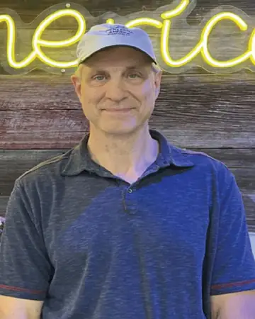 Jay Ross, smiling, in a cap and a blue polo shirt, in front of a wood-plank wall and a yellow neon sign.