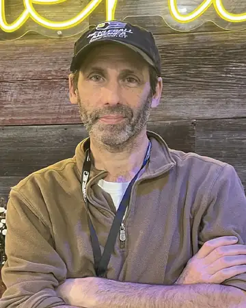 Jay Waldner, arms folded, in a Pickleball America cap and a fleece, in front of a wood-plank wall.