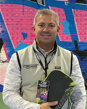 Joe Schipani, smiling, in a Pickleball America vest and lanyard, holding a paddle in front of a graffiti mural.